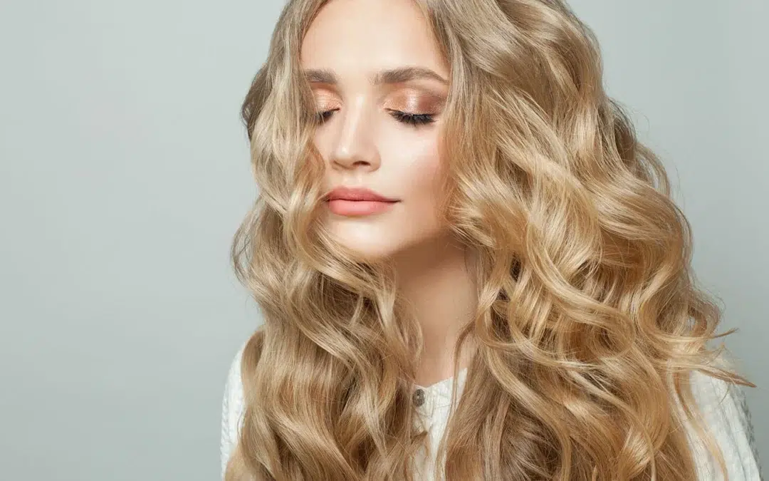 Blonde woman with eyes closed featuring long, wavy blonde hair and glowing makeup against a plain background.
